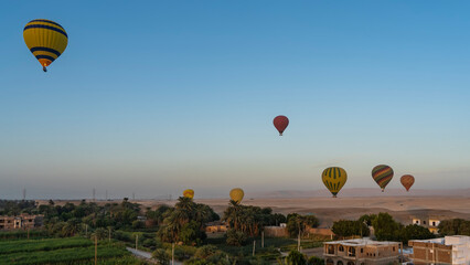Bright balloons are flying in the blue sky. Below you can see rural houses, palm trees, cultivated fields. In the distance - the sand dunes of the desert. Egypt. Luxor