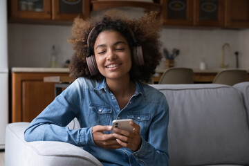 Happy serene gen Z teenage African girl in big wireless headphones listening to ambient music, relaxing tunes playlist, sitting on couch at home with closed eyes and toothy smile