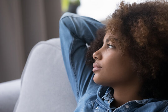 Pensive teenage African girl with natural curly hair resting on home couch, looking away in deep thoughts, thinking over problems, troubles, making serious decision