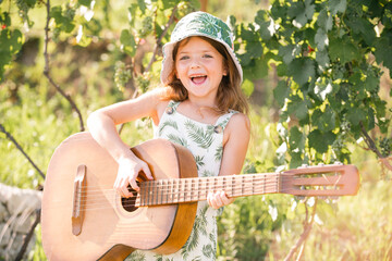 Cute child girl with blonde curly hair playing guitar on nature background. Dreamy kids face....