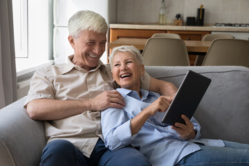 Fototapeta premium Excited older age grandparents family couple hug on couch at home use touchpad laugh watching funny video enjoy online shopping. Happy senior husband wife having fun with pad pc discuss web purchase