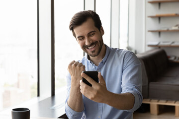 Happy excited businessman looking at smartphone screen, making winner yes gesture, smiling and...
