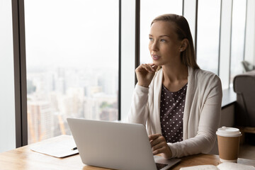 Thoughtful businesswoman pondering on project at workplace with laptop, looking out of window at...