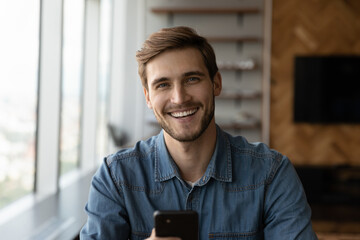 Head shot of happy young millennial man holding smartphone, looking at camera, smiling, laughing. Portrait of positive customer, smartphone user satisfied with online app, virtual service work - obrazy, fototapety, plakaty