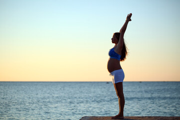 Fototapeta na wymiar pregnant girl doing yoga on the seashore in the morning against the sky and water