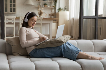 Obraz premium Focused happy student girl in big earphones watching online virtual class, workshop, webinar on laptop, studying foreign language, making video call to teacher, tutor, resting on couch
