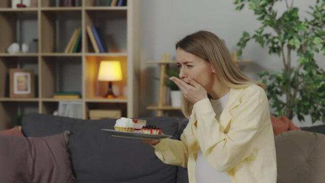 Young caucasian pregnant woman suffering from nausea when smelling cakes on plate at home. Concept of toxicosis and health problems during pregnancy time.