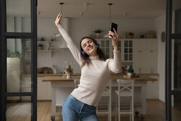 Excited carefree millennial dancer girl in wireless headphones dancing to music alone in apartment interior, listening to online playlist from smartphone, using internet media service