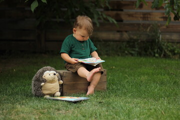 A two-year-old boy sits on a box in the park and reads a book