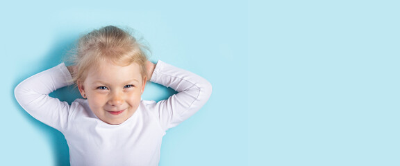 Smiling beautiful blonde girl lying down holds her hands behind her head on a blue background. Top view, flat lay. Banner