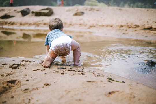 Beb&eacute; gateando hacia el agua en la playa, con las piernas y el pa&ntilde;al llenos de arena.