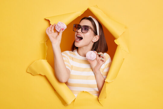 Indoor shot of hungry woman wearing striped shirt and hair band, breaks through yellow paper background, holding eating marshmallow, standing with open mouth.