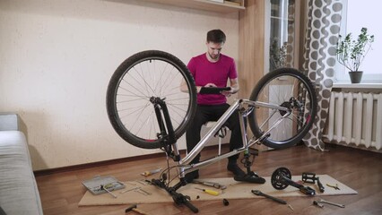 Man sits near inverted disassembled bicycle in room. Lot of parts and tools are on floor. Uncomprehending man looks at the tablet and tries to find breakdown. Guy is upset and disappointed.