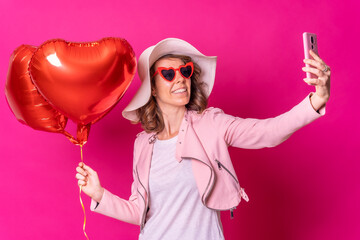 Obraz na płótnie Canvas Portrait of a caucasian woman having fun with a white hat in a nightclub with some heart balloons, taking a selfie with her mobile phone, pink background