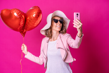 Fototapeta premium A caucasian woman having fun with a white hat in a nightclub with some heart balloons, taking a selfie with her mobile phone, pink background