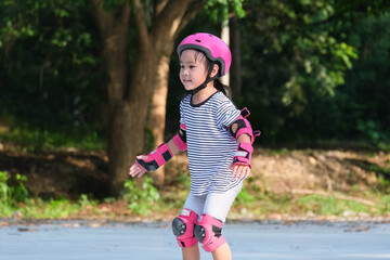 Fototapeta premium Cute Asian little girl in protective pads and safety helmet practicing roller skating in the park. Exciting outdoor activities for kids.