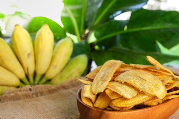 Banana Chips In Wooden Bowl On Burlap Fabric Against Banana Fruit.