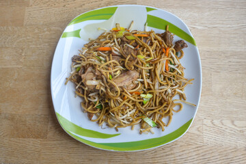 Chinese fried noodles with chicken meat, vegetables and sauce on a plate and a wooden table, copy space, high angle view from above