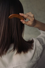 Back view of unrecognizable woman combing brown straight hair with wooden eco friendly comb while doing hair care procedure at home 