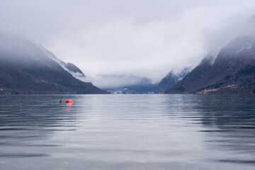 Norwegian fjord with clear water in early spring when there is still some white snow in the mountains and they are foggy, but in the dark waters of the fjord you can see a bright red buoy