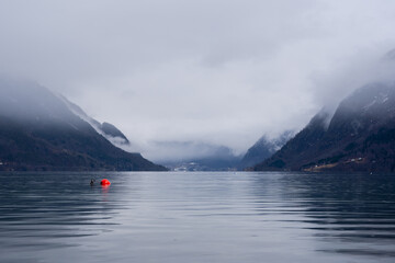 Norwegian fjord with clear water in early spring when there is still some white snow in the mountains and they are foggy, but in the dark waters of the fjord you can see a bright red buoy