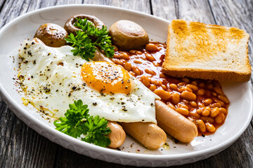 English breakfast - toast, egg, sausages, beans in tomato sauce and mushrooms on wooden table
