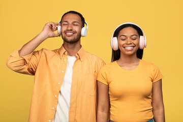 Cheerful young black couple listening to music in wireless headphones on yellow studio background