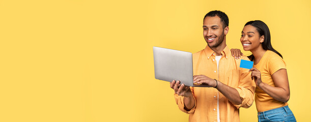 Happy young black couple holding laptop and credit card, shopping online over yellow studio background, banner design