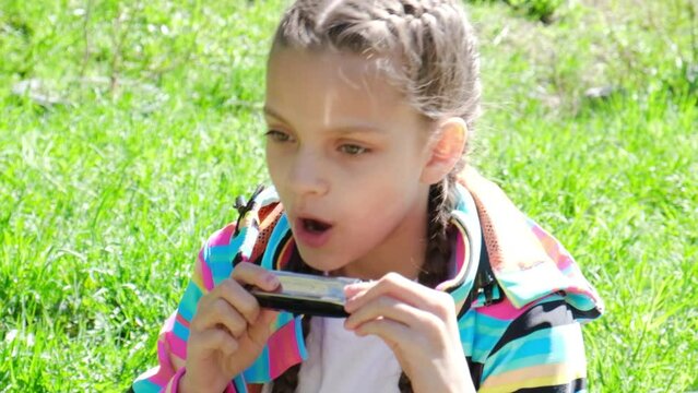 a cute teenage girl in a white T-shirt and with a hairstyle with braids plays the harmonica against the background of green grass