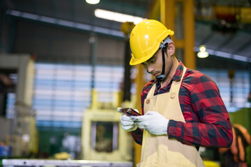 Factory worker with plaid shirt and hard hat check working order on corporate application by mobile phone in manufacturing  warehouse in night shift.
