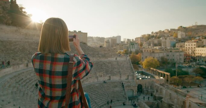 Woman takes Photo Pictures of Roman Theatre - famous landmark in Amman, capital of Jordan. Female Tourist Photography by Smartphone Jordanian or Middle East Cityscape at sunset. 4K medium orbit shot