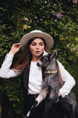 A beautiful stylish girl in a white blouse with a black vest and a white hat holds a small goat in her hands in the village. Rural life.