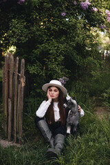 A beautiful stylish girl in a white blouse with a black vest and a white hat holds a small goat in her hands in the village. Rural life.