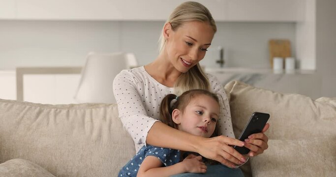 Happy young blonde mother cuddling little baby daughter, involved in using cellphone apps, communicating distantly in social network, web surfing information online or shopping in internet store.