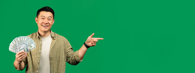 Happy middle aged asian man holding fan of money cash and pointing finger at free space, green background, panorama