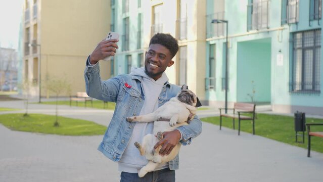 African american man with positive facial expression taking selfie photo with pug dog during walk. Cheerful young male using modern smartphone and having fun with his cute pet on fresh air.