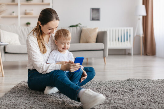 Young caucasian mother and her toddler son resting with cellphone at home, watching videos online, free space