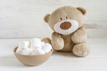 Bear toy and bowl with marshmallow on white wooden background.