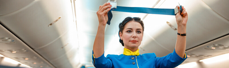 Woman stewardess in air hostess uniform holding safety belt while standing near passenger seats in...