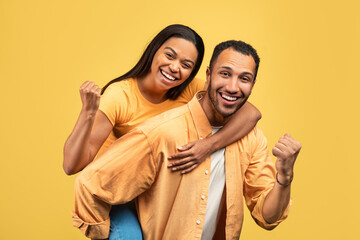 Obraz premium Attractive young African American man giving piggyback ride to his girlfriend, gesturing YES on yellow studio background