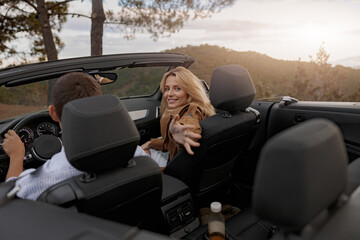 Joyful pretty woman traveling on passenger seat in cabriolet looking back holding her hand out