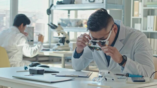 Waist up slowmo of Asian male engineer in white lab coat and special eyewear inspecting circuit board, working in laboratory with colleague