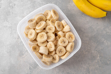 Slices of frozen banana in a container on a gray concrete background. Ingredient for smoothies, ice cream, desserts. Healthy food. Top view.