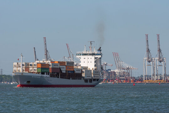 Southampton, England, UK. 2022. Small container carrier ship departing DP World, Southampton container port with backdrop of cranes and containers.