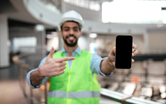 Happy millennial islamic engineer male in protective uniform, hard hat with beard point finger at phone