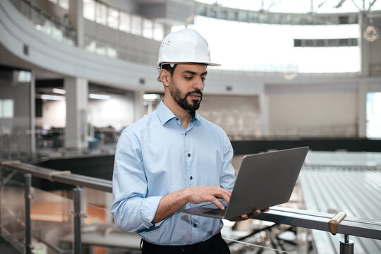 Pensive busy millennial arab guy engineer in safety helmet with beard works with computer controls factory - Powered by Adobe