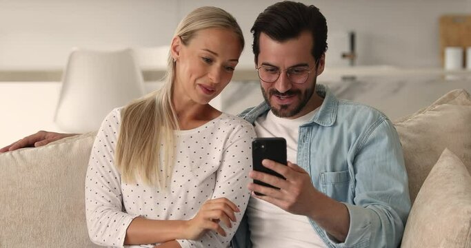 Happy bonding sincere young family couple sitting on sofa, enjoying discussing choosing goods in internet store, shopping online, watching funny photo or video content or holding web camera zoom call.
