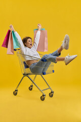 Cheerful girl holding colorful bags, while riding in shopping cart indoor. Side view of happy lady having fun with trolley, while shopping, isolated on yellow studio background. Concept of happiness.