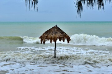 Thatch umbrella at the beach with strong waves crashing against the beach.