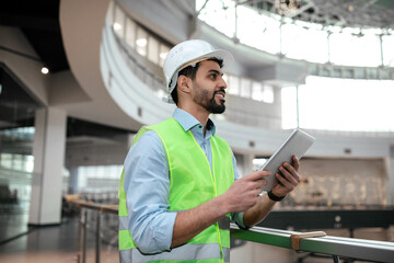 Glad millennial middle eastern man engineer in protective uniform and hardhat with beard hold tablet